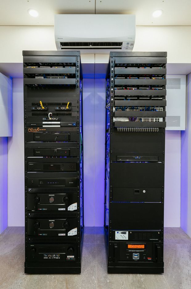 A compact technical equipment room containing two tall black server and audiovisual equipment racks, with visible patch panels, cabling, amplifiers and electronic components. A wall-mounted split-system air conditioner is installed above the racks. The room has pale walls, recessed ceiling lights, blue accent illumination between the racks, ventilation grilles, and a grey hard floor.