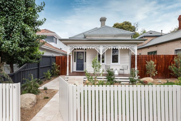 Front exterior view of a compact heritage-style single-storey home, featuring a pale weatherboard facade, hipped corrugated metal roof, ornate white decorative veranda fretwork, front steps, and a white picket fence with a landscaped garden. Neighboring homes and boundary fencing are visible on either side.