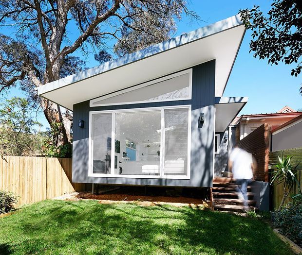 Exterior view of a compact contemporary backyard dwelling or extension with dark vertical cladding, crisp white trim, broad glazing, and a dramatic asymmetrical skillion roof. The elevated structure is accessed by a short timber stair, with a timber privacy fence, lawn, and mature trees surrounding it. A bright interior is visible through the glazing.