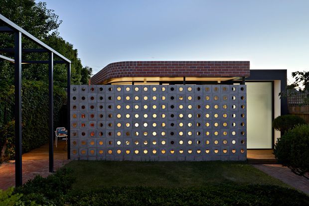 A contemporary single-storey exterior viewed from a landscaped garden. The facade features a perforated concrete breeze-block screen with circular openings, warm light glowing through the openings, dark metal framing, a curved reddish-brown brick parapet or roof form, and a partially visible glazed entry or opening. A simple dark metal pergola frames the left side, with brick paving and hedges around the building.