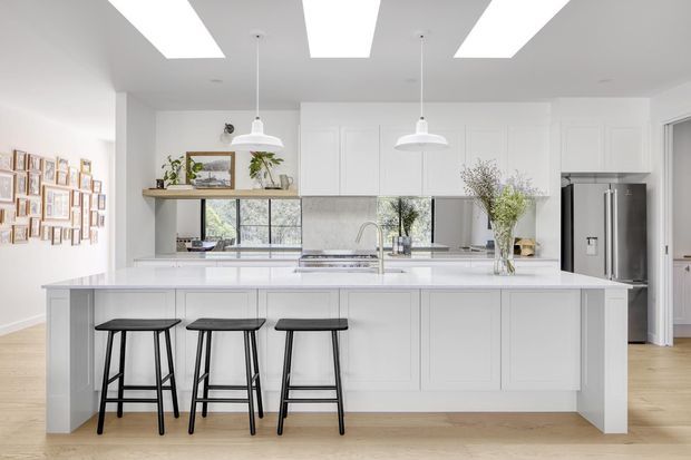 Bright, spacious contemporary white kitchen with a large island, three black bar stools, integrated cabinetry, pale stone surfaces, stainless-steel appliances, pendant lights, skylight-like ceiling openings, and light timber flooring. A framed gallery wall and open shelving add visual detail.