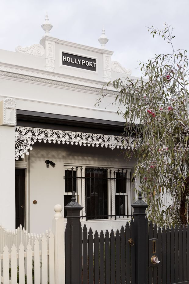 Front elevation of a refined historic terrace or townhouse facade, featuring ornate white plaster mouldings, decorative parapet urns, a sign reading “HOLLY PORT,” lace-like verandah detailing, barred windows, and contrasting black fencing and gate. A slender flowering tree partially screens the entrance.