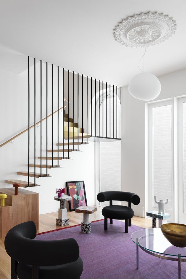 Bright, stylish living room with a sculptural open staircase featuring slim black vertical balusters and timber treads. The space has white walls and ceiling, a decorative ornate ceiling medallion, a large spherical pendant, black sculptural lounge chairs, a glass coffee table, purple carpeting, and eclectic decorative furnishings. A framed artwork and small side tables add colour and texture.