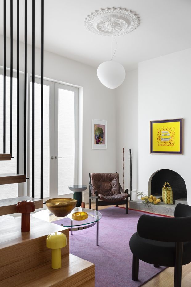 A bright, eclectic living room with a white interior, tall glazed doors and windows screened by black vertical slats, a decorative ornate ceiling medallion, and a sculptural white pendant light. The room features a black arched fireplace, purple area rug, brown leather lounge chair, rounded black armchair, glass coffee table, colourful artwork, and sculptural accent furniture in yellow, red, and wood tones.