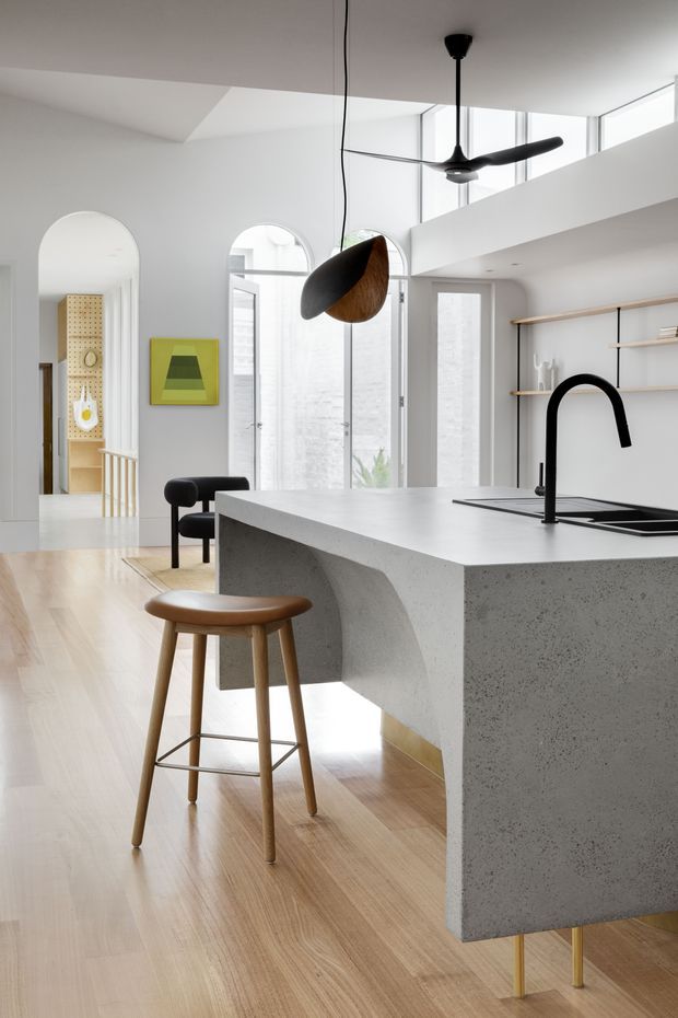Bright, refined open-plan kitchen with a large light-grey concrete island, integrated sink and sculptural black tap. Pale timber flooring, white walls, arched openings, tall arched glazing, open timber shelving, a black ceiling fan, and sculptural pendant lighting create a clean contemporary interior with mid-century-inspired furnishings.