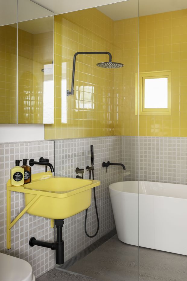 A compact, boldly coloured bathroom with glossy mustard-yellow wall tiles above a band of small light-grey mosaic tiles. The room features a bright yellow wall-mounted basin with exposed black plumbing, matte black tapware and shower fittings, a white freestanding bathtub, a mirrored wall cabinet, and a small frosted window. The floor is a smooth grey finish, and a full-height glass shower screen is visible.