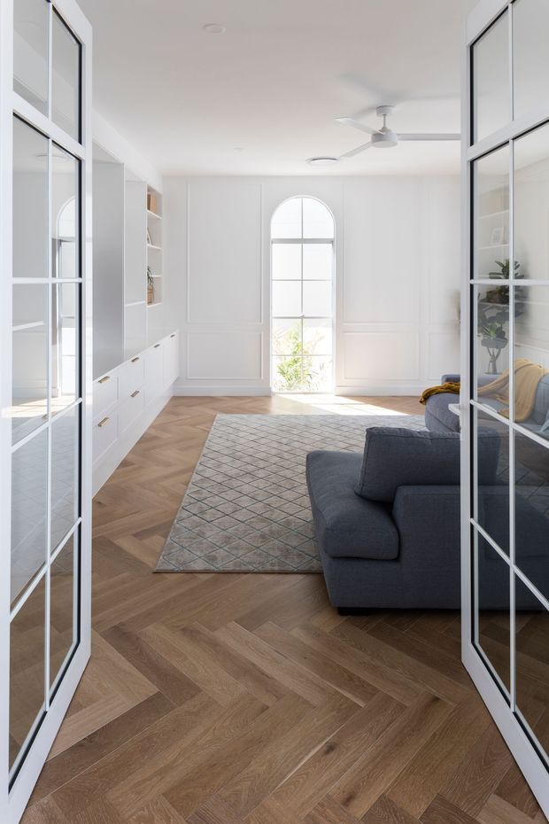 Bright, refined living room viewed through open glazed French-style doors. The room features white paneled walls, built-in white cabinetry and shelving, a tall arched window, herringbone timber flooring, a large muted geometric rug, and a dark charcoal sectional sofa. A white ceiling fan and abundant natural light create an airy contemporary-classic atmosphere.