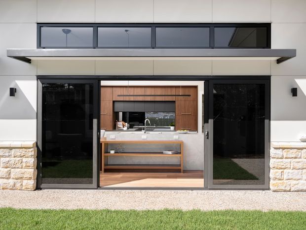 Front exterior view of a contemporary single-storey home featuring a wide covered opening with black-framed glazed sliding doors, clerestory-style upper glazing, pale rendered wall panels, and a light textured stone base. Through the open doors, a compact modern kitchen with timber cabinetry, a pale benchtop, sink, and open shelving is clearly visible. A narrow gravel strip and lawn sit in front of the facade.