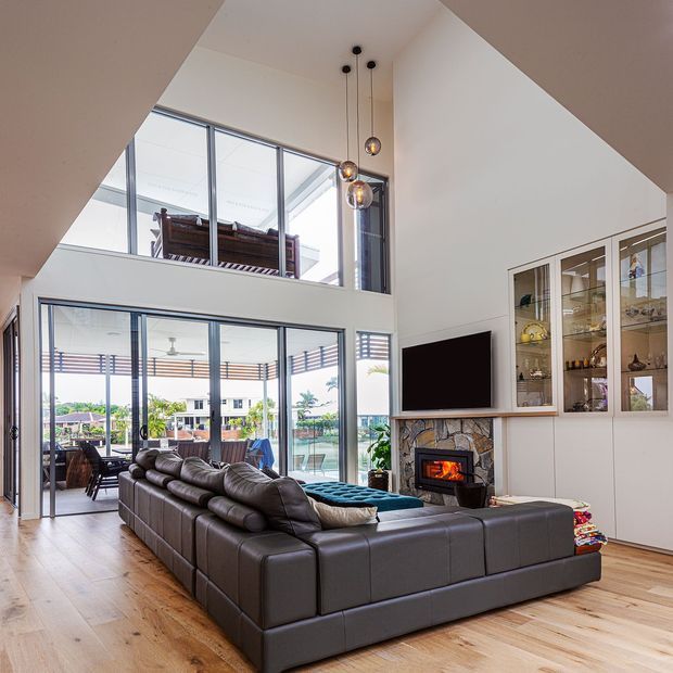 Bright double-height living room with a large dark leather sectional sofa, timber flooring, a stone-clad fireplace with built-in television, glazed display cabinetry, and extensive floor-to-ceiling glazing opening toward an outdoor entertaining area. Pendant lights hang in the tall void, with an upper-level glazed area visible above.