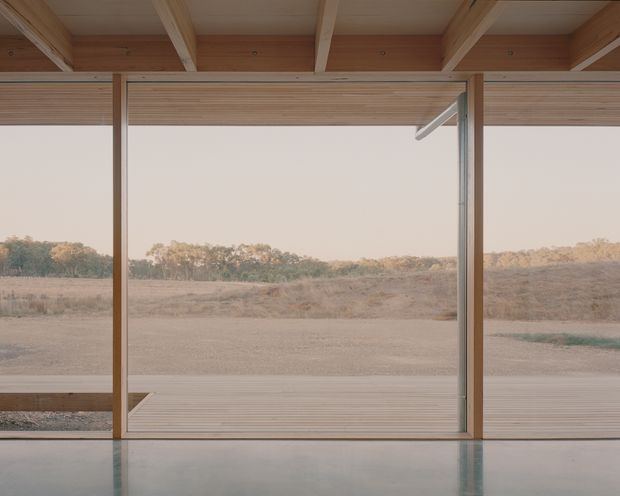 A minimalist interior opening onto a broad rural landscape through full-height glazed sliding doors. The space features exposed light timber ceiling beams and a timber-lined ceiling, slim dark window and door frames, a polished smooth grey floor, and a timber deck outside. The composition is quiet and highly open, with strong indoor-outdoor connection and simple geometric lines.