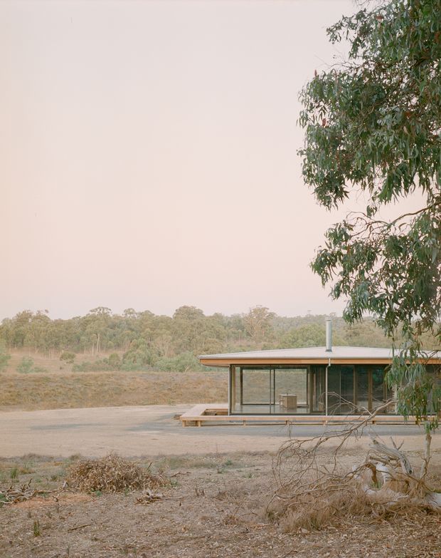 A low, single-storey modern pavilion set within a dry, open bushland landscape. The building has extensive floor-to-ceiling glazing, slender dark window frames, a shallow metal roof with broad eaves, and a small raised timber deck or platform. A simple chimney or flue projects through the roof. Eucalyptus trees and sparse, dry ground surround the structure, creating a quiet rural Australian setting.