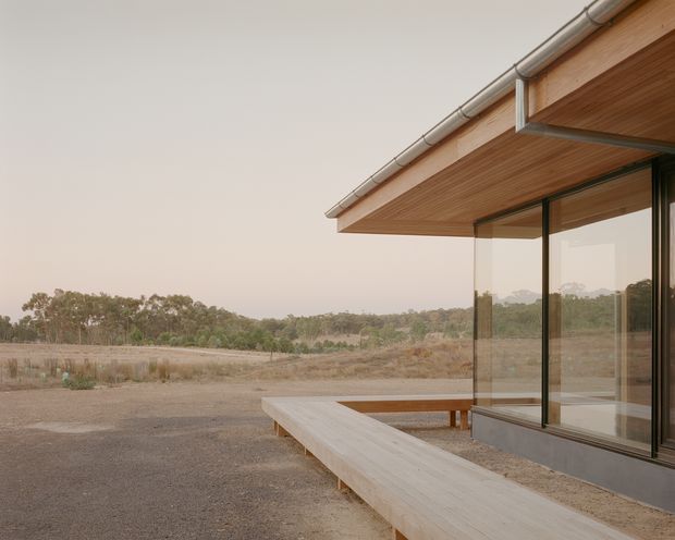 A minimalist single-storey building exterior viewed from a broad paved or compacted outdoor area. The composition focuses on a projecting timber-lined roof soffit, slim dark-framed floor-to-ceiling glazing, and a long pale timber bench or deck-like platform along the facade. The building sits in a sparse rural landscape with low vegetation and trees in the distance, creating a restrained contemporary architectural scene.