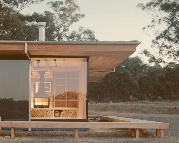 A compact single-storey modern pavilion set within a wooded landscape. The building has a broad shallow roof with a pronounced timber soffit and projecting eave, extensive floor-to-ceiling glazing, dark framed window panels, and a raised timber deck or bench along the front. Through the glazing, a wood-burning stove and neatly stacked firewood are visible. The restrained natural palette and connection to the surrounding bush give the image a calm, contemporary cabin-like character.