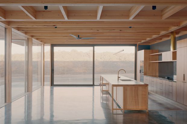Open-plan contemporary kitchen and living area with extensive floor-to-ceiling glazing overlooking a dry, mountainous landscape. The room features a polished concrete floor, extensive warm timber ceiling beams and lining, timber kitchen cabinetry, a white island benchtop, integrated shelving, and dark-framed sliding glass doors.