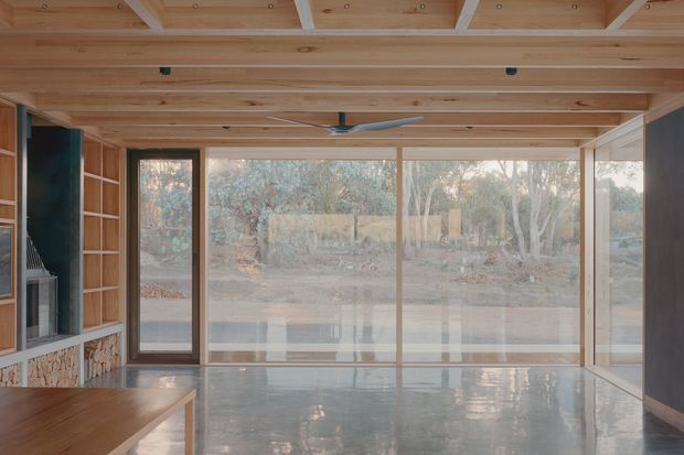 A timber-lined open-plan interior with exposed structural beams and a warm wood ceiling, broad floor-to-ceiling glazing, and a polished concrete floor. Built-in timber shelving and a stone-faced fireplace are visible at the left, with a timber dining or work surface in the foreground. The glazing overlooks a wooded outdoor setting, and a ceiling fan hangs centrally.