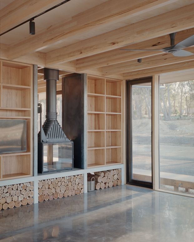 A warm, timber-lined living room with exposed wood ceiling beams, full-height built-in shelving, and a central black wood-burning fireplace. Firewood logs are neatly stacked in open storage beneath the shelving. Large glazed doors and windows bring in views of the surrounding trees, while a ceiling fan and track lighting complete the functional interior. The polished grey concrete floor reflects the natural light.