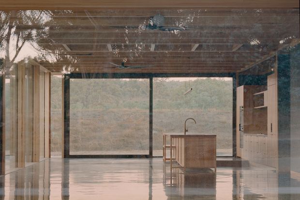 A serene, open-plan kitchen viewed from inside a highly glazed modern home. The room features a central timber island with integrated seating and a curved faucet, extensive floor-to-ceiling glazing, dark slender frames, timber-lined ceiling, pale cabinetry, and highly reflective polished flooring. A wooded landscape is visible beyond the glass.