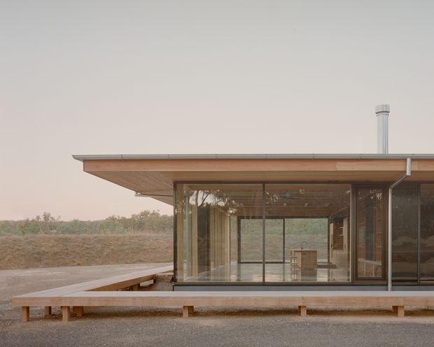 A low-profile single-storey modern pavilion set in a dry rural landscape. The building features a broad shallow roof with exposed warm timber soffits, extensive floor-to-ceiling glazing, slim dark window frames, and a raised timber deck wrapping around the front. The minimalist interior is visible through the glass, with a compact kitchen or joinery element and sparse furnishings. The muted earth-toned setting and strong horizontal lines create a calm, refined architectural composition.
