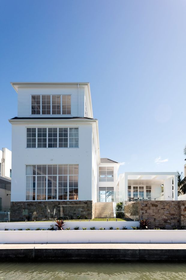 A bright exterior view of a large two-storey white coastal-style residence beside a canal or waterfront. The home has extensive white-framed multi-pane glazing, flat and low-pitched roof forms, a covered open terrace, stone retaining walls, landscaped planting, and a waterfront edge in the foreground.
