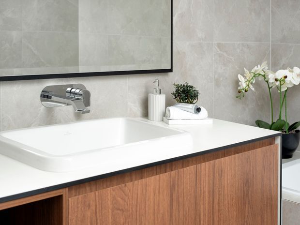 Close-up view of a refined bathroom vanity featuring a large white integrated basin and white benchtop, warm timber-grain cabinetry, a wall-mounted chrome tap, pale stone-look wall tiles, and a framed mirror. Small toiletries and a potted orchid add restrained decorative detail.