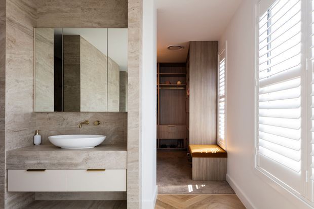 A refined bathroom and adjoining walk-in wardrobe are shown. The bathroom features full-height beige stone-look wall and floor surfaces, a floating white vanity with integrated stone benchtop, white vessel basin, brushed brass wall-mounted tapware, and mirrored cabinetry. Through the opening is a timber-toned walk-in wardrobe with open shelving, hanging storage and a built-in window seat. Bright natural light enters through white plantation shutters.