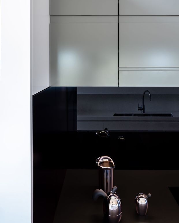 A close-up view of a dark, highly refined kitchen. Glossy black cabinetry and benchtops contrast with pale grey upper cabinetry, while a dark sink and curved black tap sit against a charcoal splashback. Chrome kitchen hardware and a polished metallic kettle or coffee set appear in the foreground.