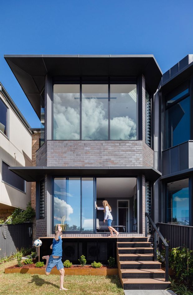 Exterior photograph of a narrow multi-storey contemporary townhouse, featuring dark metal cladding and trim, large floor-to-ceiling glazing, exposed light brickwork, projecting flat roof and balcony-like slab edges. A timber stair leads from the lawn to an open lower-level terrace, with children playing in the foreground. Bright blue sky and neighbouring urban residences frame the building.