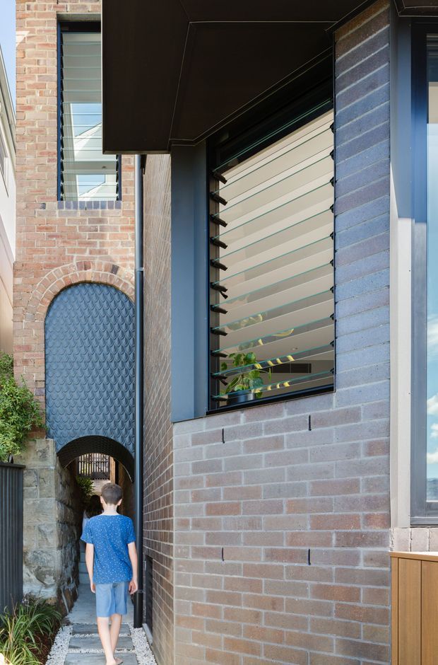 Vertical exterior architectural photograph of a narrow residential passage beside a contemporary brick house. The composition features pale and blue-grey brickwork, a projecting dark soffit, a high window with horizontal louvres, and a distinctive arched pedestrian opening with a dark patterned metal gate. A person walks through the passage, with planting and stonework at the lower left.