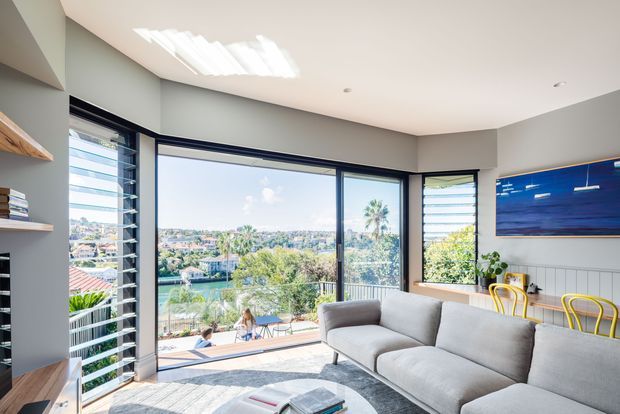 Bright contemporary living room with expansive corner glazing and louvred windows overlooking a leafy suburban waterfront setting. The room features a large light-grey sectional sofa, built-in open shelving, dark-framed windows and doors, a pale wood or concrete-toned floor, white walls, and a dining area with yellow chairs. A large blue artwork provides a strong accent. The high windows and open layout create an airy, relaxed interior.