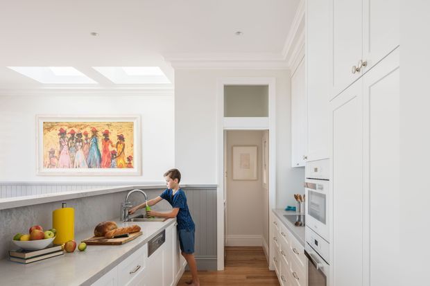 Bright, refined white kitchen with extensive paneled cabinetry, a long grey stone island, integrated double ovens, timber flooring, and a child at the sink. The room features decorative wall panelling, high skirting and cornice details, skylights, and colourful artwork, creating a spacious traditional family-kitchen setting.