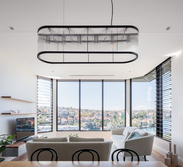 Bright contemporary living room viewed from behind a dining table, with expansive corner glazing overlooking a dense residential waterfront or urban landscape. The room features a large neutral sectional sofa, a television on low joinery, timber flooring, white walls and ceiling, timber floating shelves, venetian-style horizontal blinds, and a prominent elongated black-framed pendant light with a suspended linear cage form.