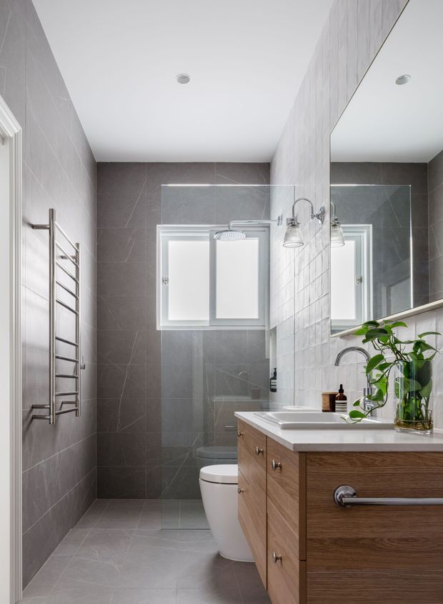 A compact contemporary bathroom with large grey tiled walls and floor, a frameless glass shower enclosure, white toilet, wall-mounted towel warmer, and a timber vanity with a white integrated basin and chrome fixtures. A large mirror, frosted window, recessed ceiling lights, and a potted plant complete the bright, practical space.
