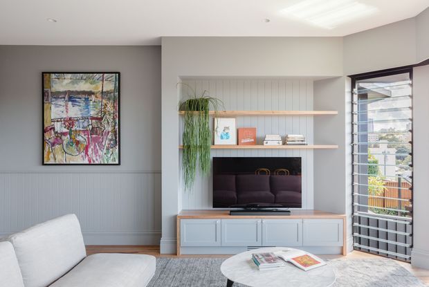 Bright contemporary living room with pale grey vertical wall panelling, a built-in entertainment unit and open shelving around a television, framed abstract artwork, indoor greenery, a white sofa, round coffee table and large louvred glass door opening to an exterior balcony or terrace. A skylight adds natural illumination.