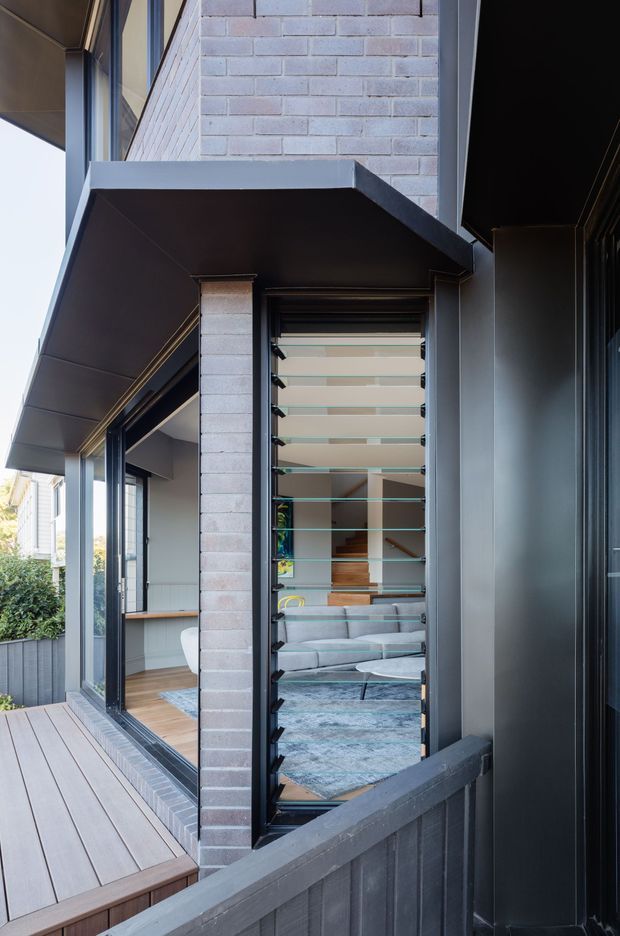 Exterior architectural photograph of a contemporary multi-level residential building, viewed along a narrow timber-clad deck or balcony. The facade combines pale exposed brick, dark metal cladding and framing, large glazed openings, and a prominent louvred window or screen. Through the opening, a furnished living room with a sofa, rug, and timber stair is visible.