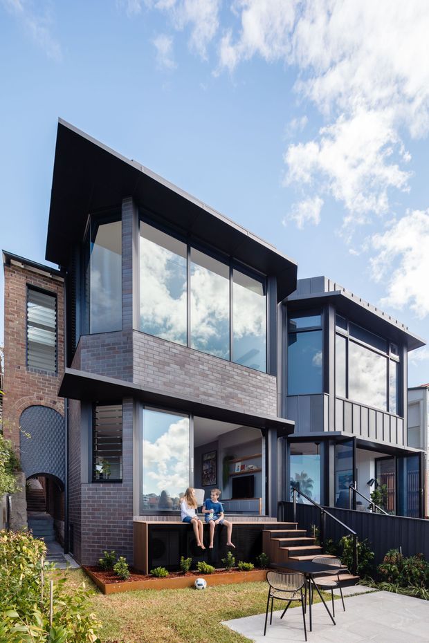 Exterior view of a contemporary multi-storey urban residence with dark brick and charcoal cladding, strong angular roof planes, extensive floor-to-ceiling glazing, and projecting box-like volumes. A landscaped backyard includes a paved patio, garden beds, stairs, and built-in outdoor seating. The image also shows interior living space through the glazing.