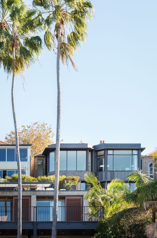 A bright exterior view of a contemporary multi-storey residential building framed by tall palm trees. The facade combines extensive floor-to-ceiling glazing, dark metal roof and framing, exposed brickwork, balconies with black metal balustrades, and timber-toned louvred shutters. Clear blue sky and landscaped planting create a sunny coastal or suburban atmosphere.