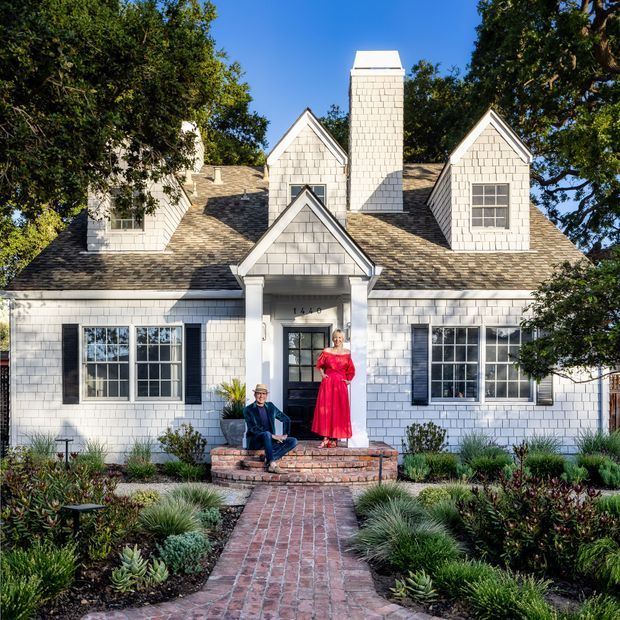 Front exterior view of a symmetrical, shingle-clad suburban house with multiple steep gables, a prominent central chimney, dark shutters, and a covered entry porch. A brick path and brick steps lead through a landscaped garden to the front door.