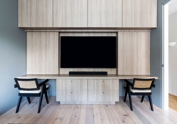 A compact, carefully fitted media and work area with pale timber built-in cabinetry spanning the wall, a recessed flat-screen television, and a low central console. Two black-framed chairs with woven seats sit at either side, suggesting a shared workspace or occasional seating. The room has pale timber plank flooring, cool grey-blue walls, and a bright doorway opening to an adjacent space.