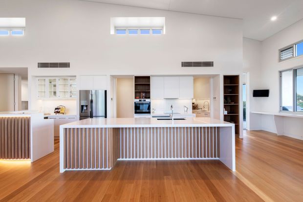 Bright open-plan kitchen with a large central island featuring vertical timber slat detailing, white cabinetry, integrated appliances, stainless-steel refrigerator, and a pale stone benchtop. Warm timber flooring, high ceilings, clerestory windows, extensive glazing, and built-in shelving create an airy contemporary interior connected to an adjacent living area.