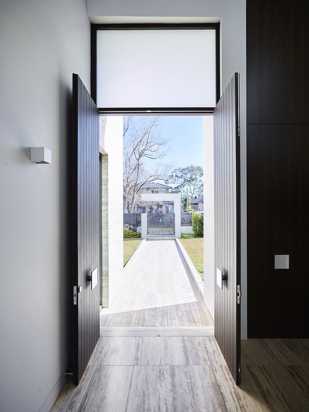 A contemporary residential entryway viewed from inside, with tall dark ribbed double doors opened toward a paved front path and gate. The interior features pale stone-look flooring, white walls, dark timber cabinetry or wall panelling, minimalist square wall fixtures, and a large glazed transom above the doorway. Bright daylight and a suburban garden are visible outside.