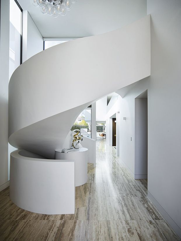 Bright minimalist interior hallway dominated by a sculptural, curved white staircase or stair enclosure. The space features a long polished stone or marble-look floor, white walls, tall glazing, recessed doorways, and a contemporary glass-bubble chandelier. A small console and decorative sculpture sit beneath the stair.