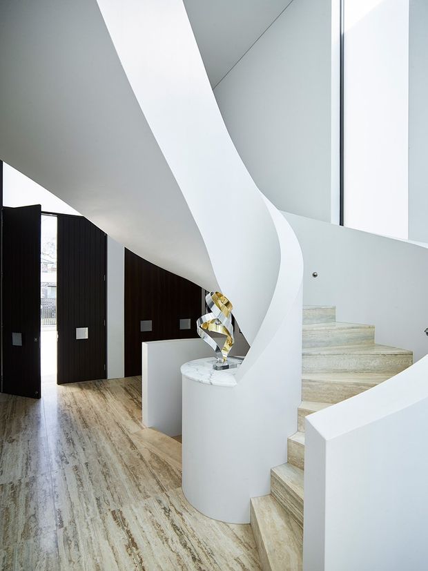 A sculptural interior stairway with a sweeping white curved balustrade and sculpted central form. Pale stone steps rise beside dark timber pivot doors and wall panels, while light stone flooring, white surfaces, and tall glazing create a bright, refined contemporary entry space. A small sculptural object sits on a rounded white built-in pedestal.