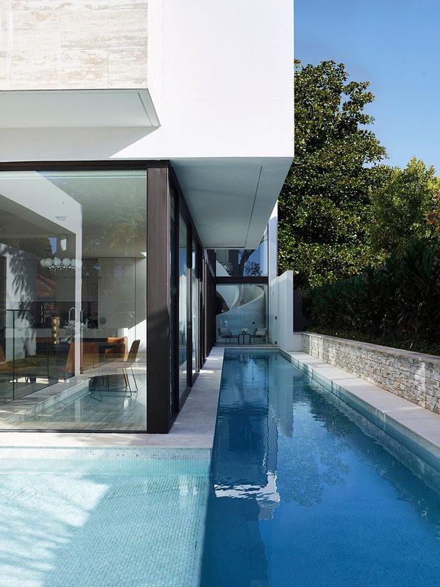 A contemporary multi-level residence with crisp white rendered volumes, dark-framed floor-to-ceiling glazing, and strong cantilevered forms. The view focuses on a long narrow swimming pool running alongside the house, with a shallow tiled section in the foreground, a pale stone retaining wall, and landscaped greenery beyond. The glazing reveals a refined dining and living area inside, while the pool reflects the building's geometric architecture.