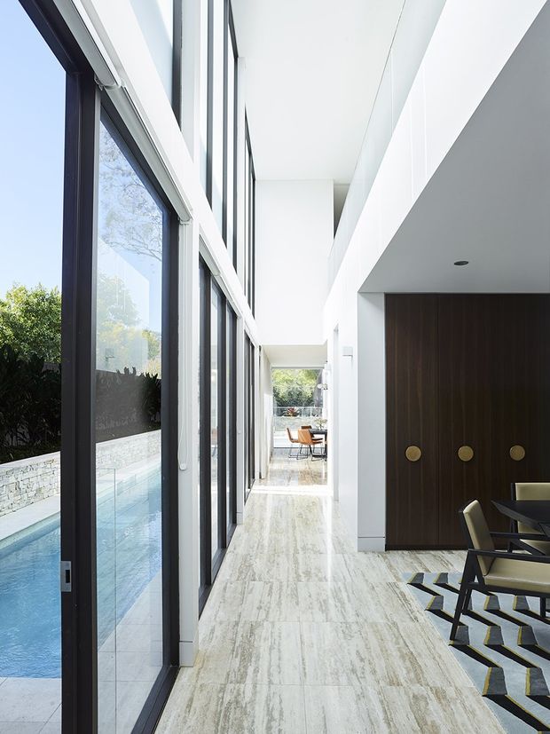 A bright, narrow contemporary interior corridor lined with full-height black-framed glazing overlooking a swimming pool and landscaped garden. The corridor has a pale stone floor, white walls and ceiling, and a dark timber built-in wall with circular brass-toned pulls. A dining area with dark-framed furniture and a patterned rug is partially visible at right, while another outdoor seating area appears through the glazed passage.