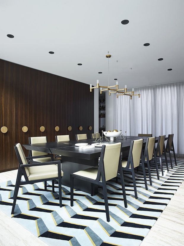 A sophisticated dining room with a long dark dining table surrounded by upholstered mid-century-inspired armchairs. The room features a dramatic dark timber wall with circular brass accents, a white ceiling with recessed downlights, a sculptural brass linear chandelier, full-height sheer curtains, and a bold geometric rug in black, white, grey-blue, and yellow. The overall design is refined, high-contrast, and contemporary with mid-century influences.