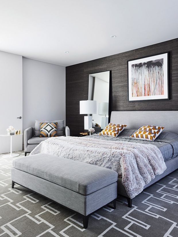 A contemporary bedroom with a large upholstered grey bed, matching upholstered bench, geometric grey-and-white rug, and grey armchair. A dark textured feature wall behind the bed includes a tall mirror and framed abstract artwork, while patterned mustard, white, and grey cushions add colour. The room has a white ceiling with recessed downlights and a minimal white side table.