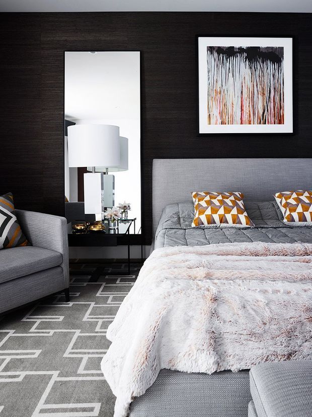 A contemporary bedroom with a dark timber feature wall, upholstered grey bed and armchair, geometric patterned rug and cushions, a fluffy white throw, bedside table, large mirror, and abstract wall artwork. The room has a layered, monochromatic palette with orange geometric accents.