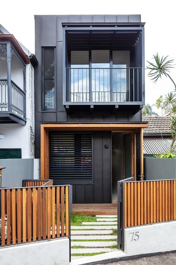 Front exterior view of a narrow, two-storey urban townhouse with a dark metal-clad facade, projecting upper-level balcony, timber-framed entry, timber slat fencing, and a stepped concrete-and-grass pathway. Neighbouring traditional homes are visible at the sides, while the main dwelling has a crisp contemporary composition with strong vertical and horizontal lines.