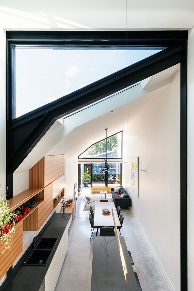 Bright, narrow open-plan interior viewed from an elevated position, combining a contemporary kitchen, dining area and living room beneath a dramatic angled ceiling and large black-framed triangular clerestory glazing. The kitchen has warm timber joinery, a dark benchtop and island, while a dining table and compact living area extend toward full-height glazing at the rear. Polished concrete flooring, white walls and abundant daylight create a clean, airy feel.