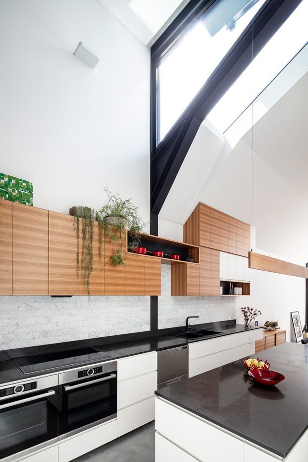 A bright, modern kitchen with a long wall of warm timber cabinetry, white marble-look tiled splashback, black stone benchtops, integrated stainless-steel appliances, and a large central island. A dramatic angled skylight or clerestory window rises above the kitchen, with indoor trailing plants adding greenery. The kitchen has a tall, open, contemporary architectural character.